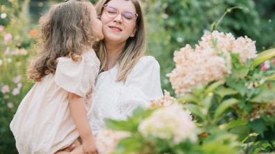 Une jeune fille embrasse tendrement sa mère sur la joue tout en étant portée par elle, entourée de fleurs colorées et de verdure luxuriante en plein air.