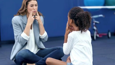 Image d’une praticienne de la psychomotricité assise par terre face à une enfant et montrant une expression faciale avec ses deux mains