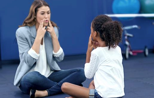 Image d’une praticienne de la psychomotricité assise par terre face à une enfant et montrant une expression faciale avec ses deux mains