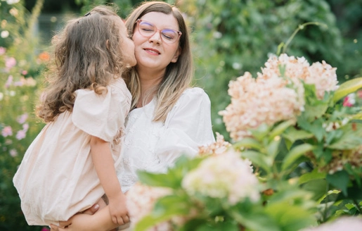 Une jeune fille embrasse tendrement sa mère sur la joue tout en étant portée par elle, entourée de fleurs colorées et de verdure luxuriante en plein air.